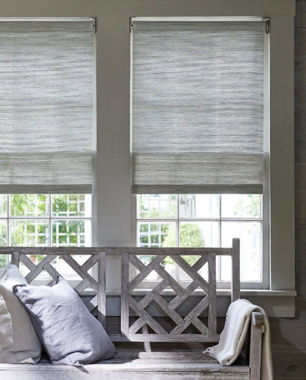 A wooden bench sits in front of two windows covered with motorized Woven Wood Shades in silver grassweave material.
