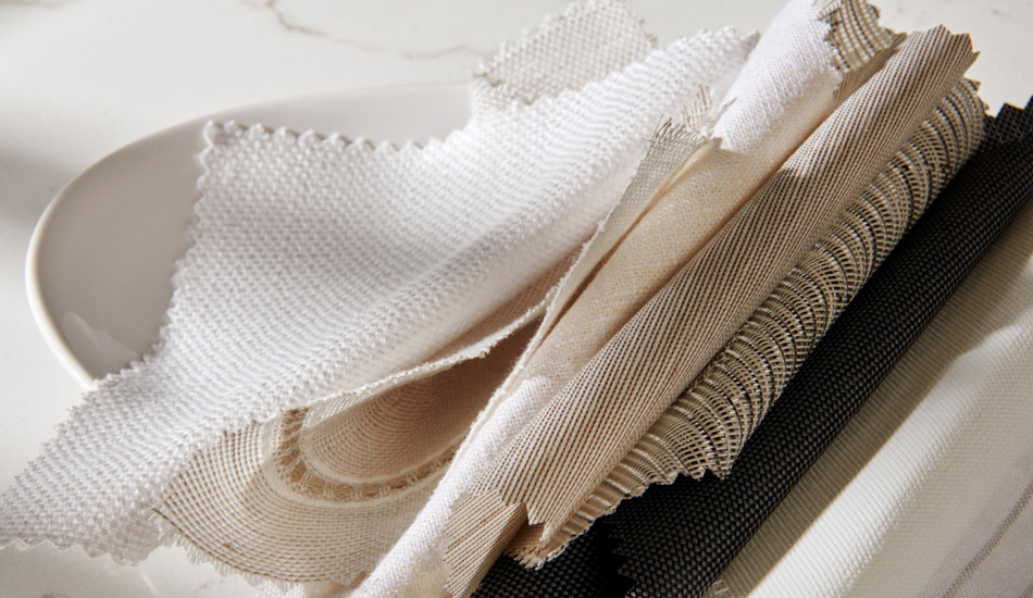 Various fabric swatches from The Shade Store stacked one over the other in a white bowl on a white table top